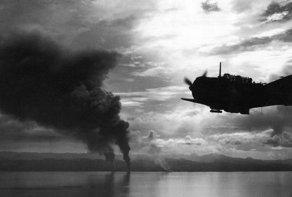A military aircraft is seen in flight with smoke rising from bombs dropped on islands in the distance