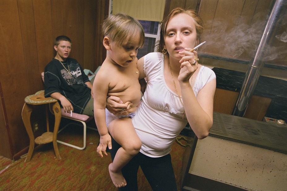 A young woman smoking  holds a baby with a young man in the background