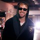 The actor Ryan Gosling smiles, wearing black sunglasses and a hood over his head