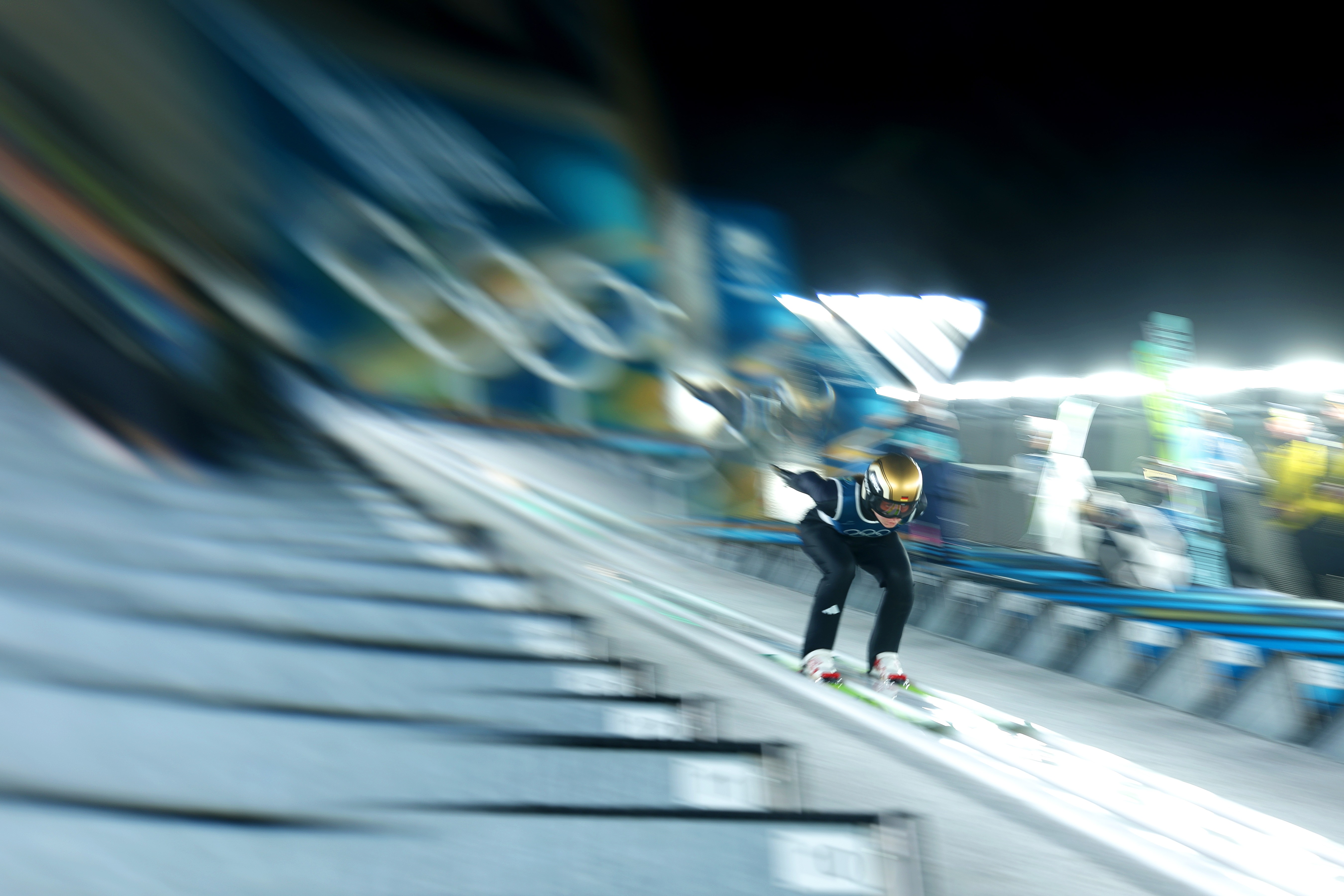A motion-blurred image of a ski jumper at the start of their jump.
