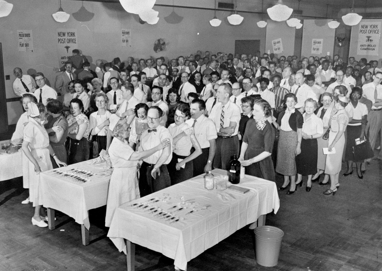 Dozens of adults, fill a room, standing in lines, awaiting vaccine shots from several nurses.