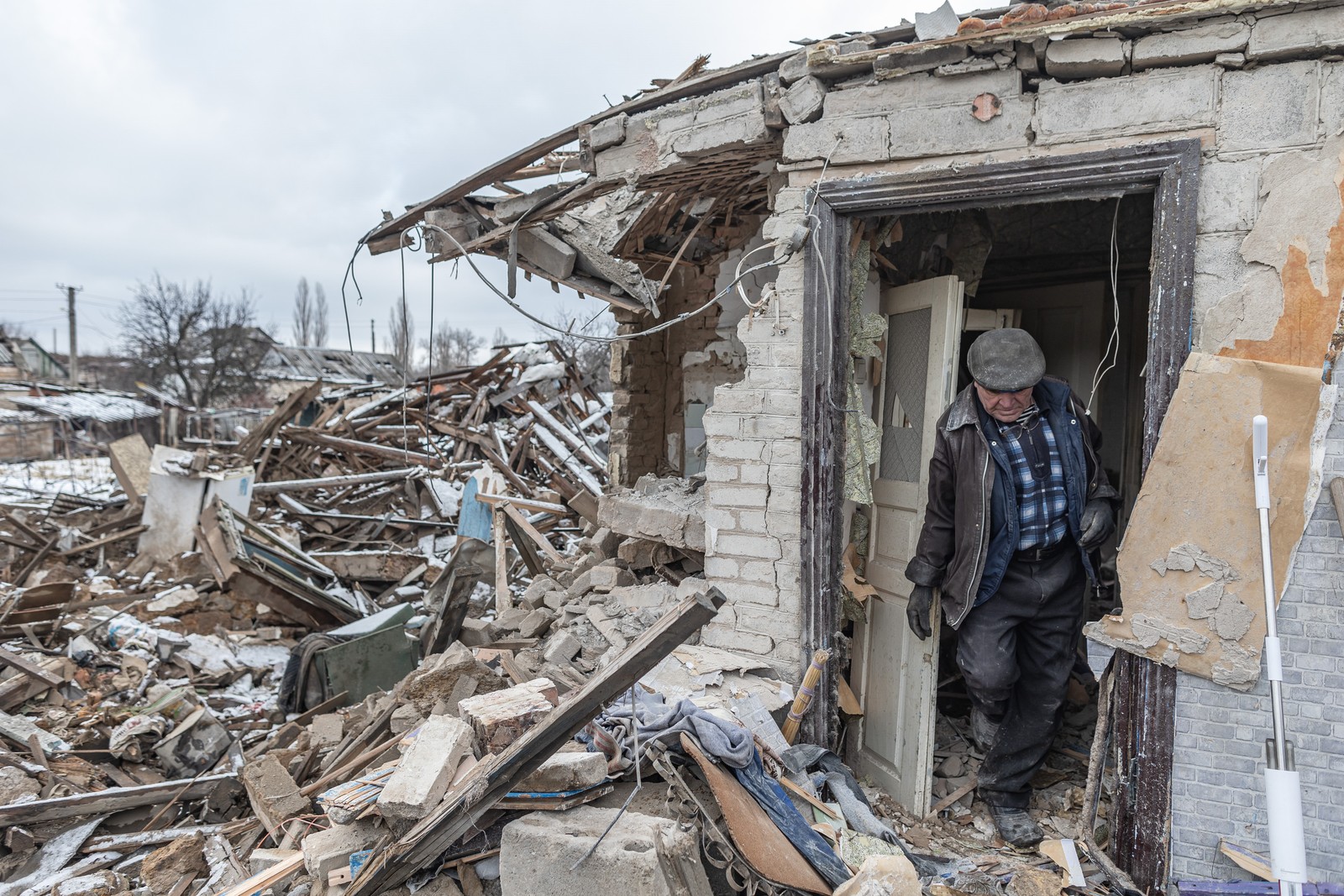 A man walks through the doorway of a heavily-damaged house, standing beside huge piles of debris.