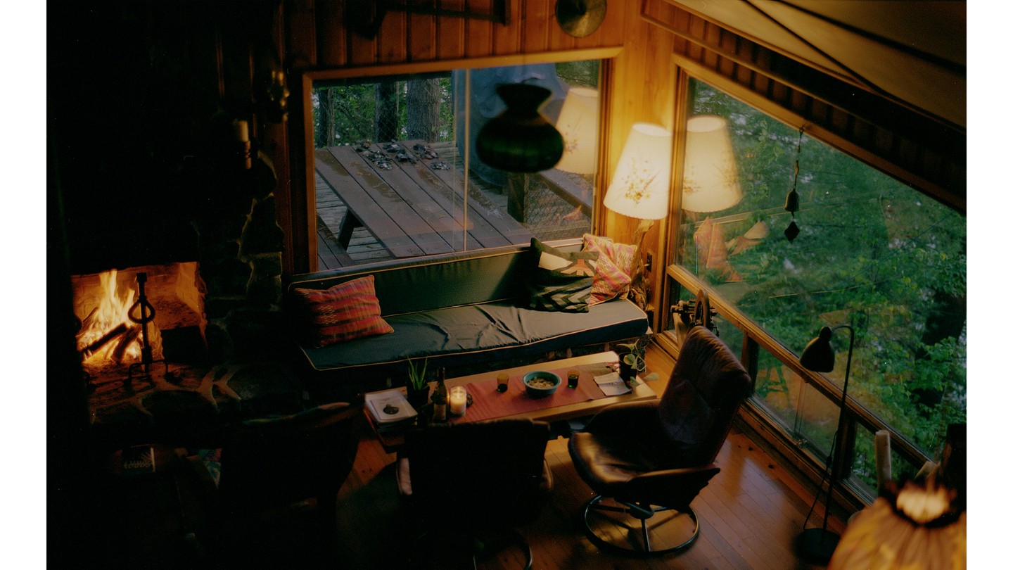 A color photograph of an living room at dusk, am empty leather chair, a couch with fabric cushions and a roaring fireplace