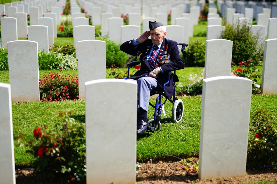 An elderly veteran wearing many medals sits in a wheelchair, saluting, among many rows of headstones in a cemetery.