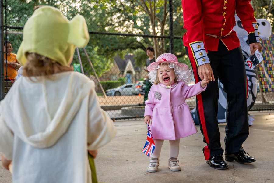 Një fëmijë i vogël me një kostum të Halloween-it të Mbretëreshës Elizabeth qan ndërsa mban dorën e një të rrituri të veshur si roje pallati.