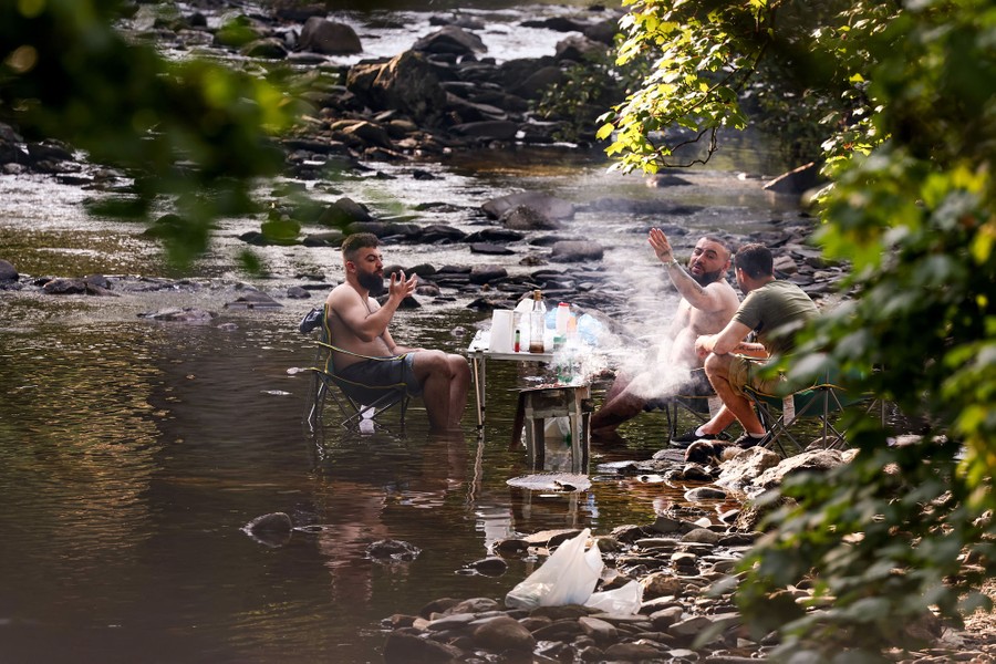Several people sit in chairs near a table and barbecue, all set up in a shallow stream.