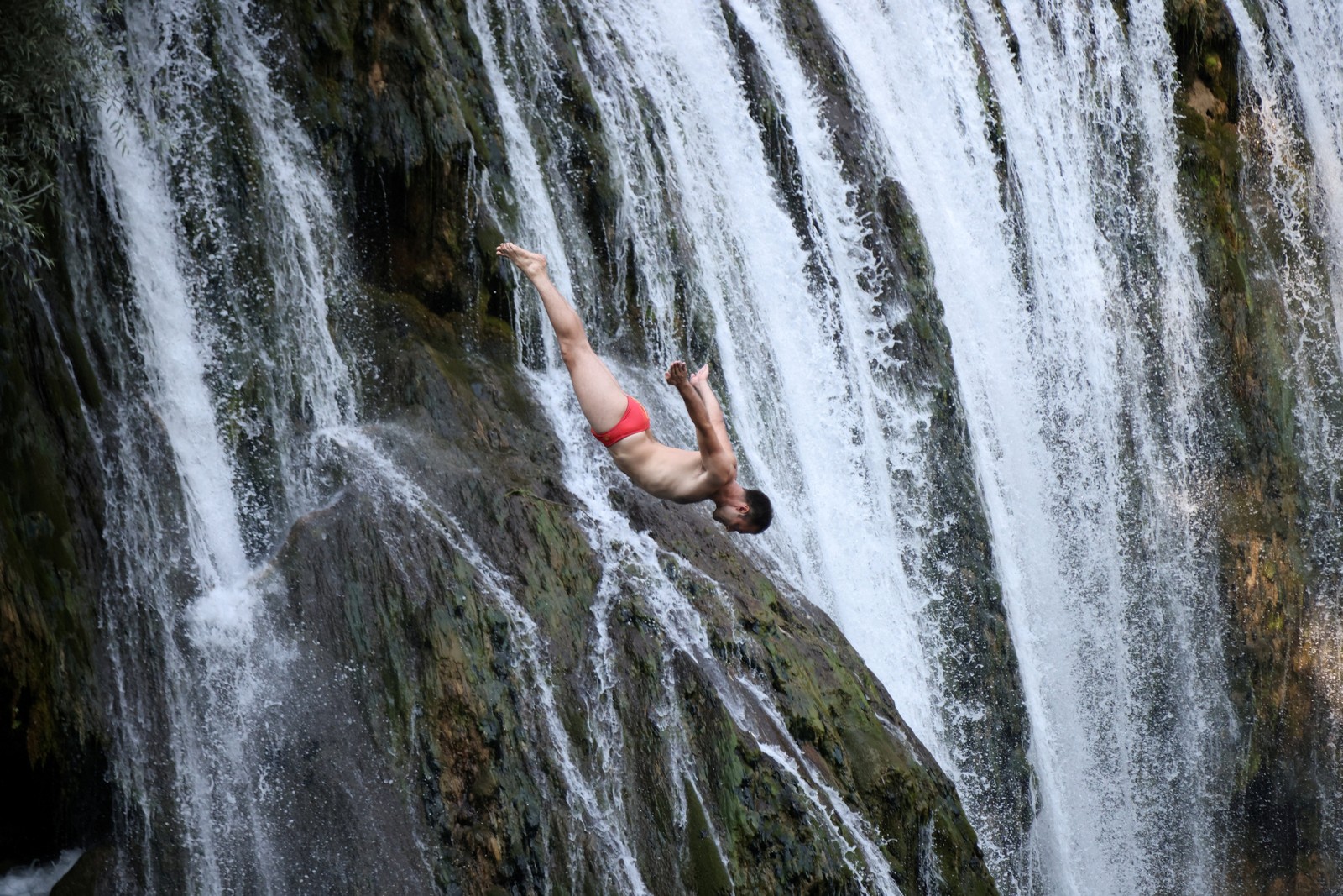 A diver plunges, falling past a waterfall.