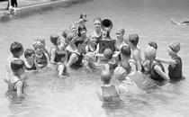 Children listen to a battery-powered radio receiver that is set on a table in a swimming pool.