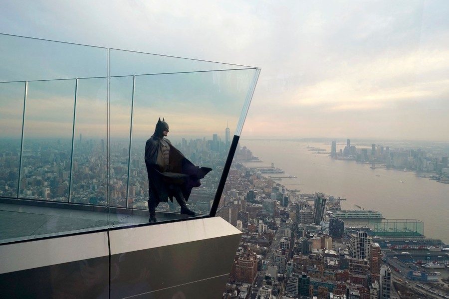 A person dressed as Batman stands on a high sky deck above the New York skyline.