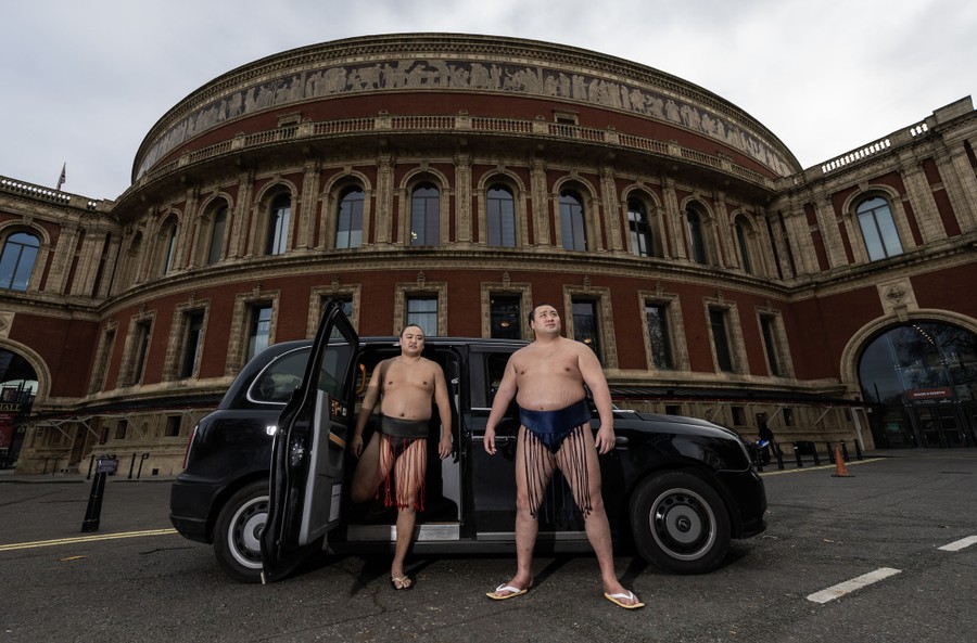 Two sumo wrestlers, wearing their traditional wrestling gear, stand outside a black taxi in a London street.