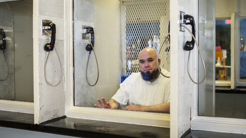 A photograph of a bearded man behind prison glass and surrounded by black phone receivers; his fingertips touch the window