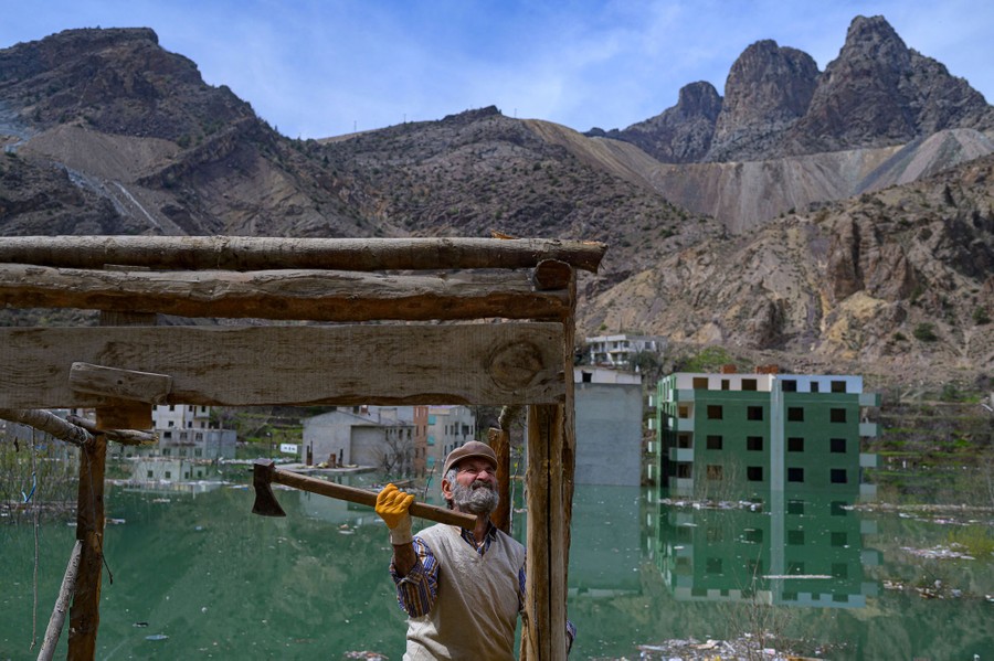 A man uses an axe to tear down a small wooden structure beside a reservoir full of flooded buildings.