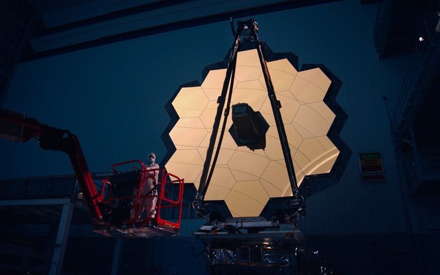 A photograph of the James Webb Space Telescope's primary mirror—an array of hexagonal, gold-covered tiles—illuminated in a dark room