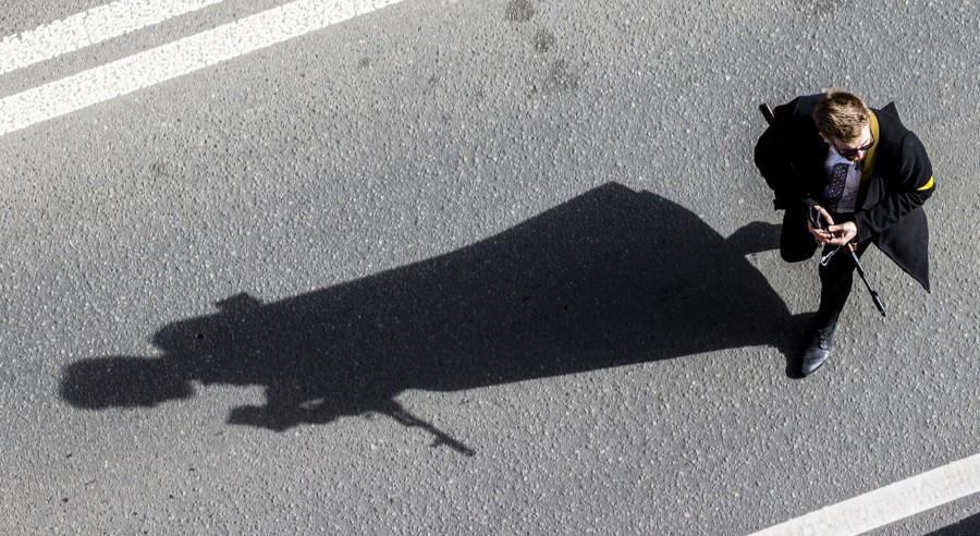 A young man wearing a civilian coat stands in a street with a rifle, casting a long shadow.