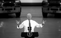 A black-and-white photo of Joe Biden speaking at a podium in front of two trucks.
