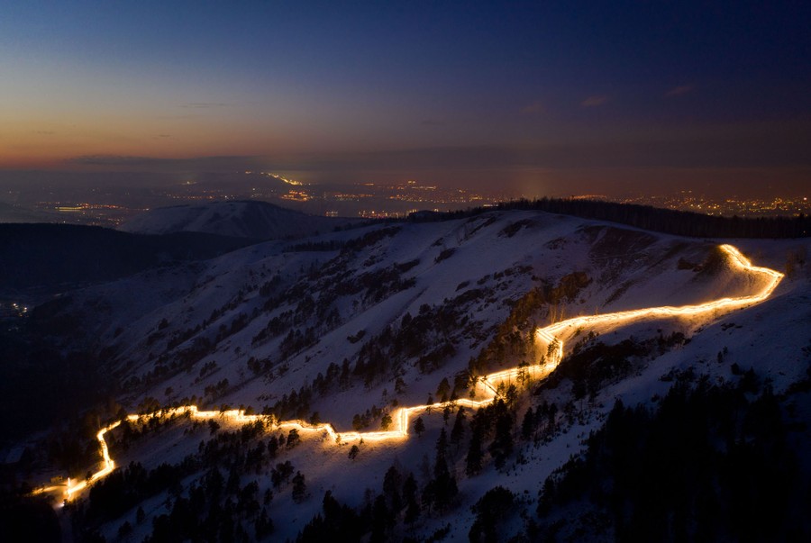 A view looking down on a mountainside that has a long, illuminated, zig-zag pathway cutting across it.