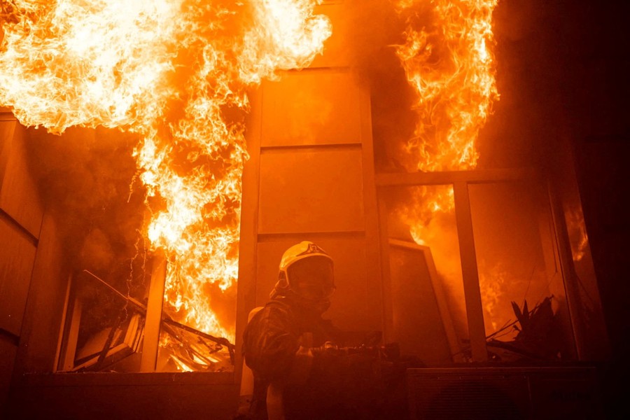 A firefighter works, standing beside a building where flames erupt through two large windows.
