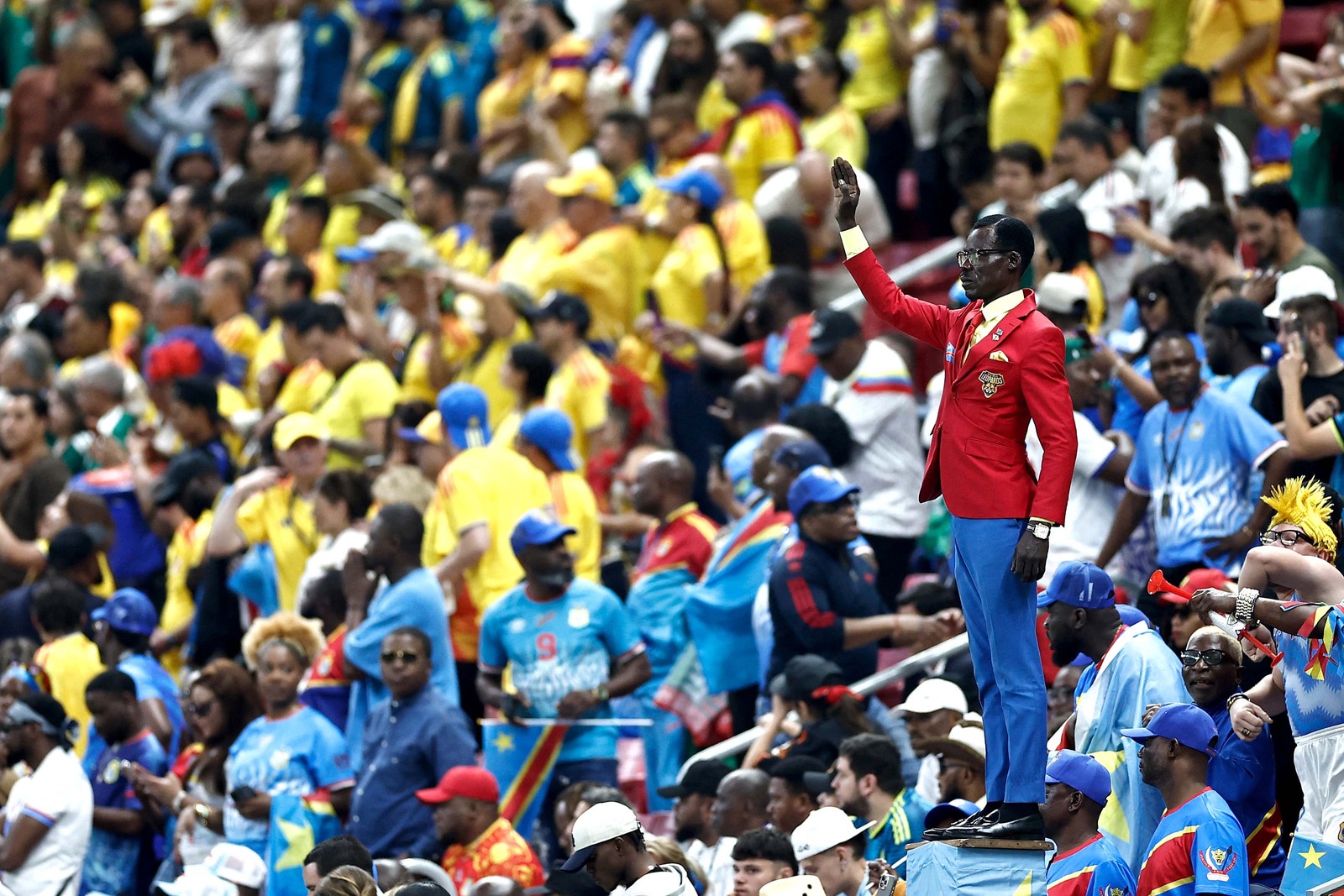 A football supporter stands stiil, bolt-upright, one arm raised, among a stadium crowd during a game.