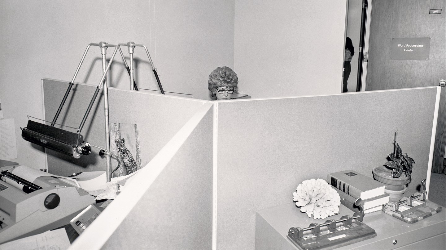 black and white photo of office cubicles with a female office worker's head visible in the back over the cubicle walls