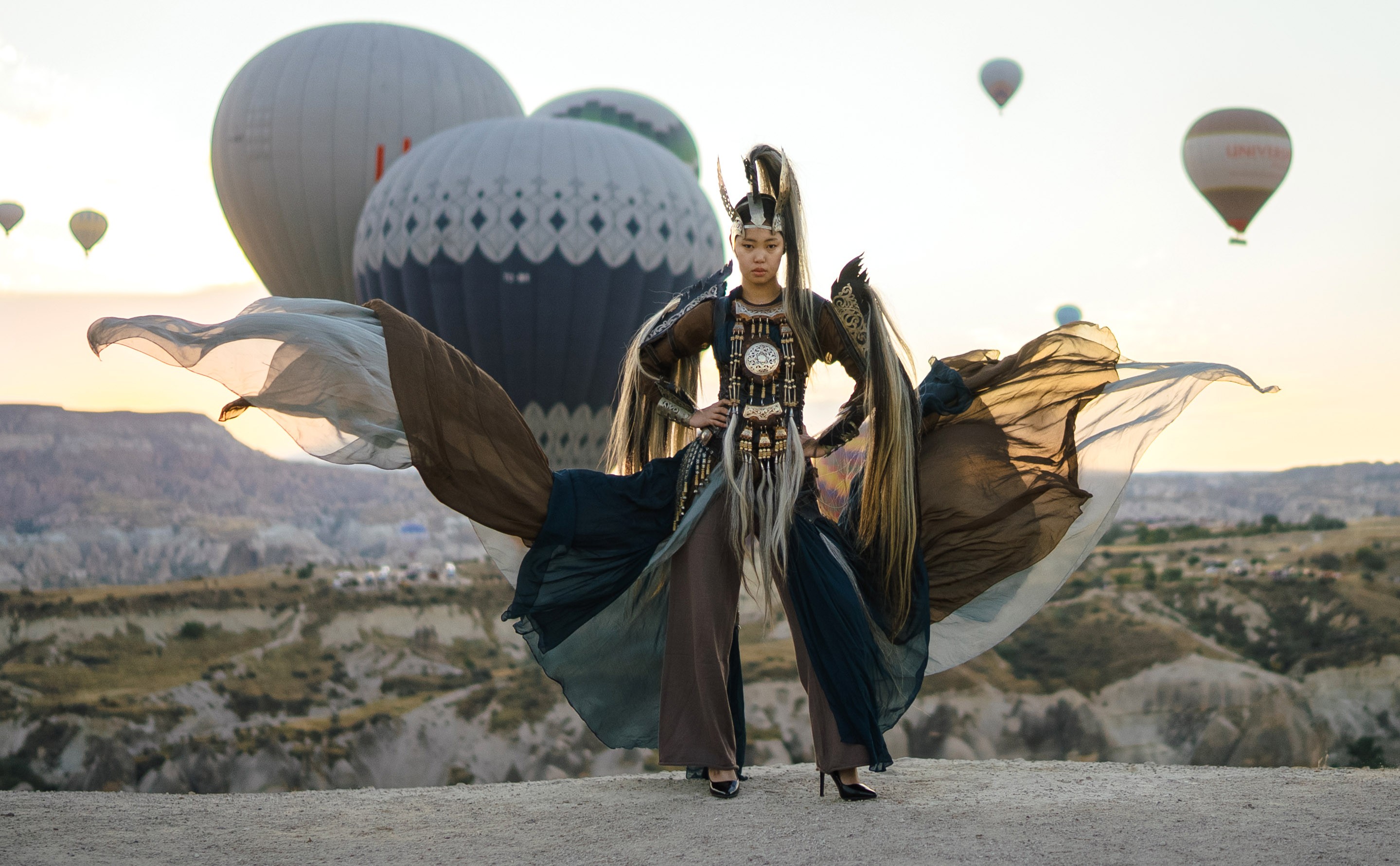 A model poses outside, in front of a half-dozen passing hot-air balloons, wearing a flowing garment based on traditional designs.