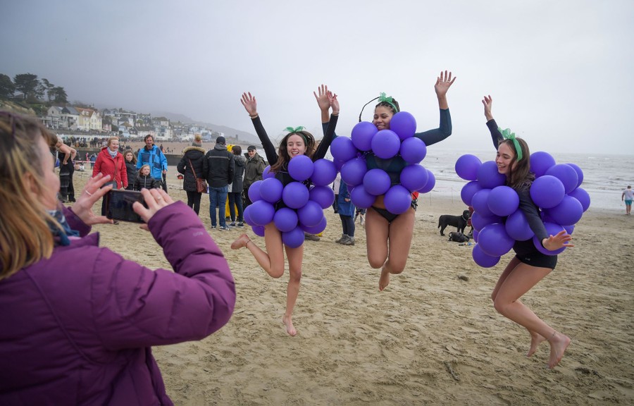 Three girls dressed as grapes (purple balloons clustered on their bodies) jump for their friend, who is taking a photo, on a beach.