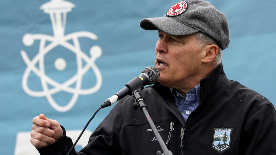 Washington Governor Jay Inslee speaks into a microphone in front of a banner showing an atom and the Seattle Space Needle.