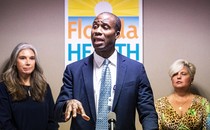 Florida Surgeon General Joseph Ladapo delivers a speech in a blue suit as two women watch behind him.