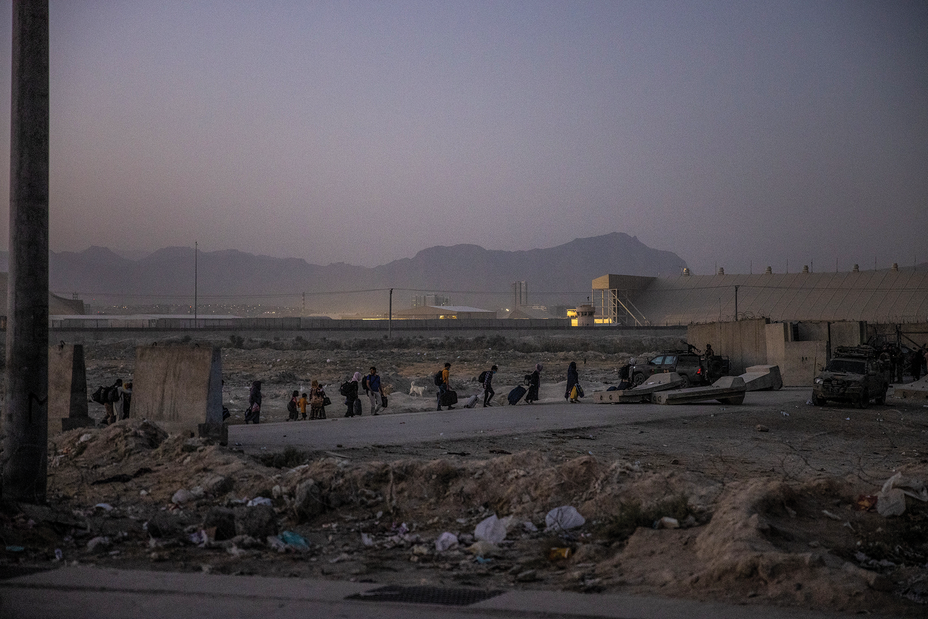 photo: a line of figures in a debris-strewn area outside the walled airport with mountains in background in dim hazy light