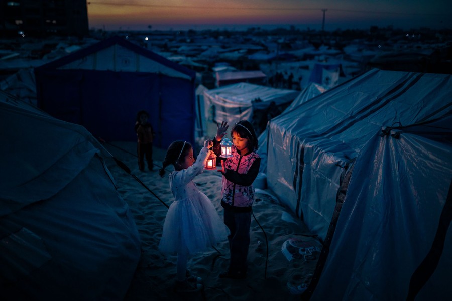 Two children hold decorative lanterns while standing outside tents in a refugee shelter.