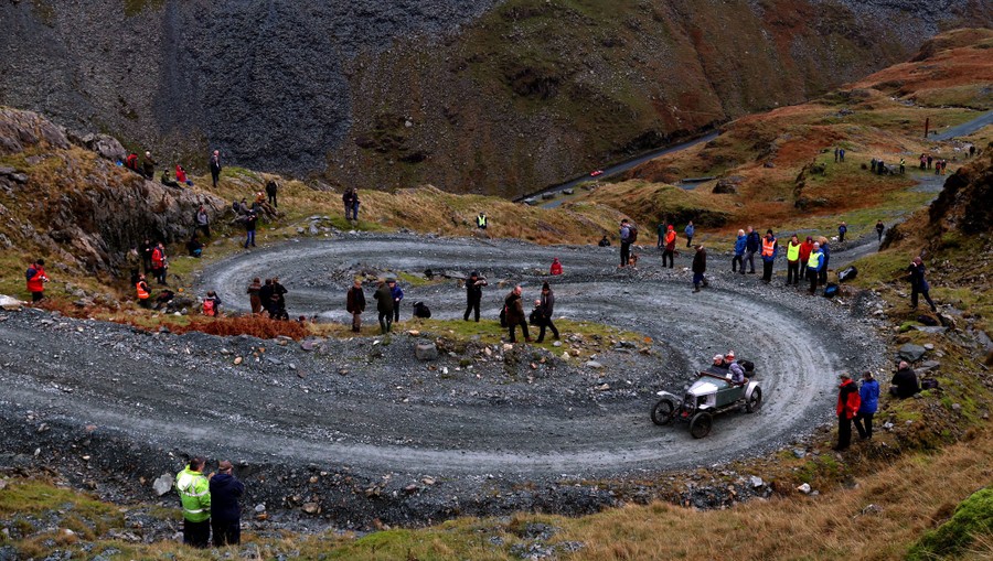 People drive a vintage car up a twisty dirt road, watched by onlookers.