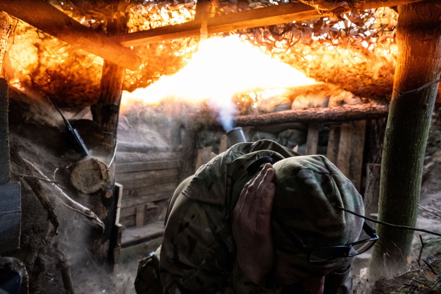 A soldier kneels in a trench, covering their ears as a mortar fires behind them.