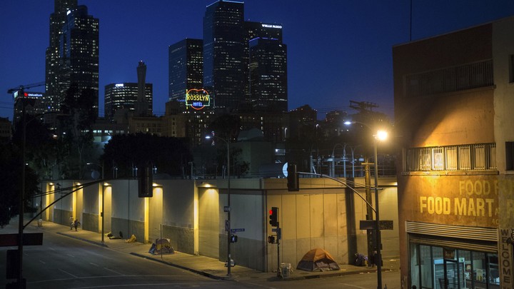 Homeless people sleeping in tents under streetlamps with the LA skyline beyond.