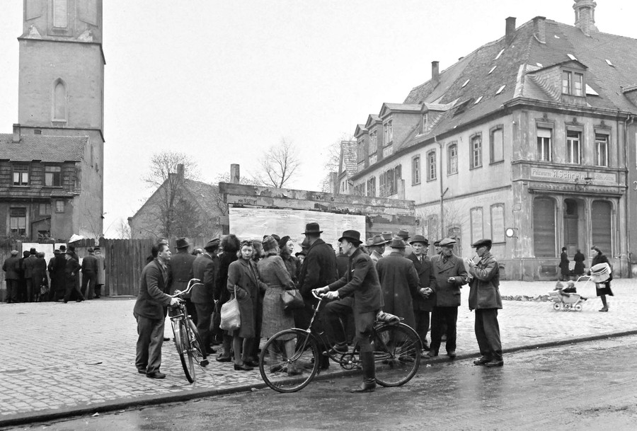 Two clusters of townspeople in a small German town gather in squares, reading posted notices in 1945.