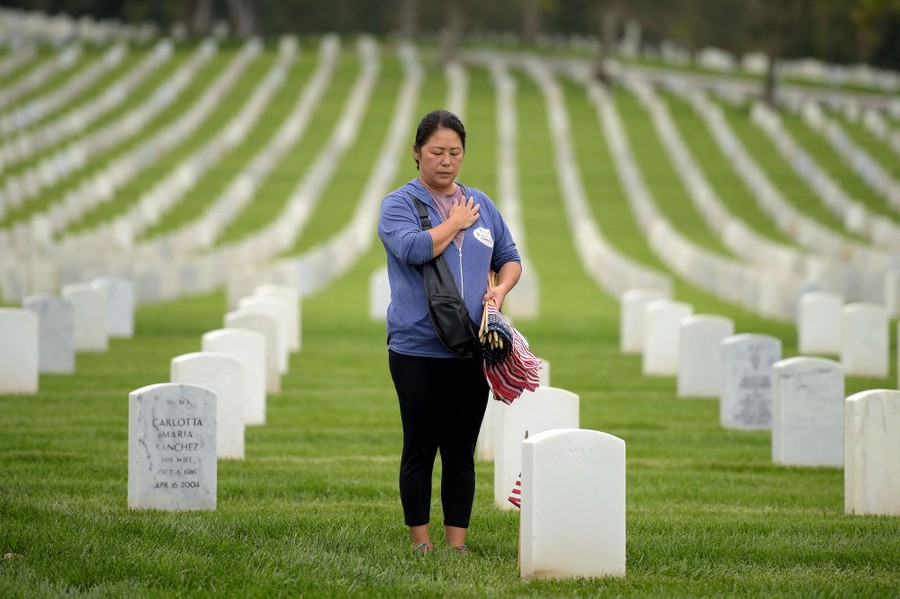 A woman stands among hundreds of headstones in a military cemetery, holding one hand over her heart, her other hand full of small American flags.