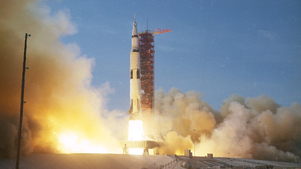 The Saturn V Rocket begins to launch, leaving behind it a trail of fire and clouds of smoke.