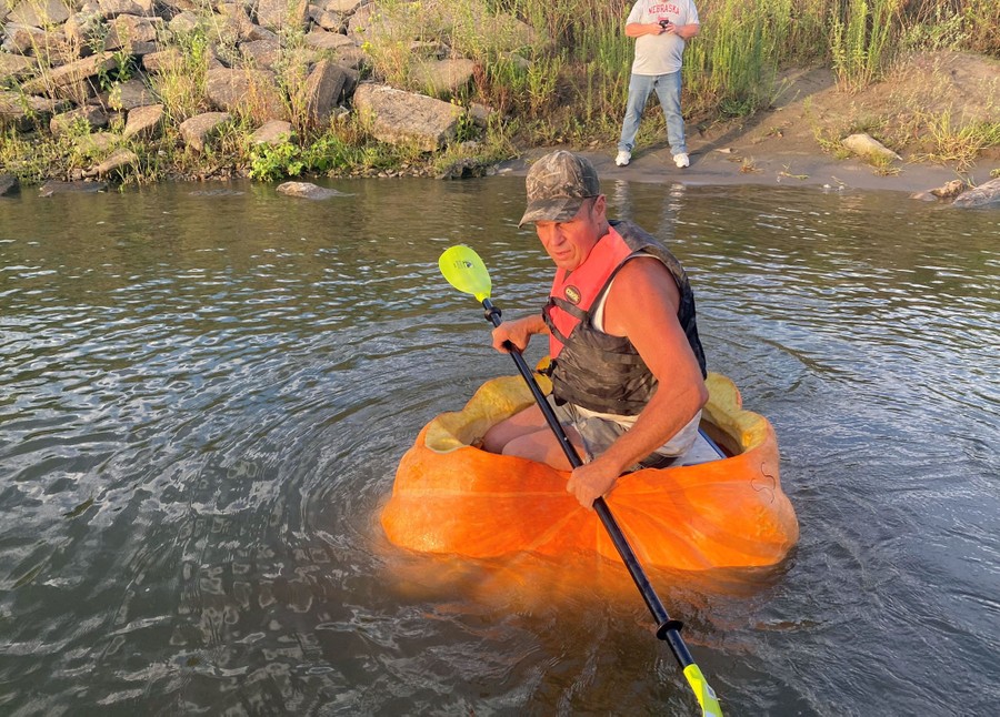A man sits in a large hollowed-out pumpkin, floating in a river, holding a paddle.