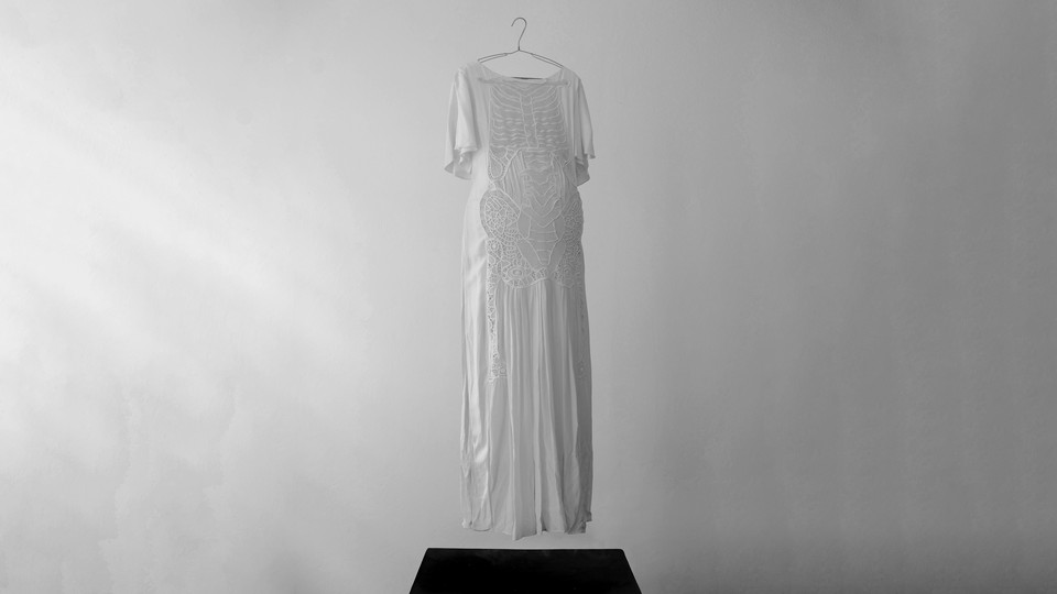 A black and white photograph of a floating white floor length dress with faint embroidering of a skeleton and a slight belly