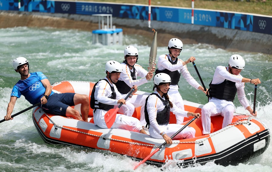 Five people paddle and steer an inflatable raft, while another sitting in the raft holds an Olympic torch.