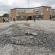 Kids play on a broken basketball court. The net on the hoop is missing. A brick school is in the distance. 