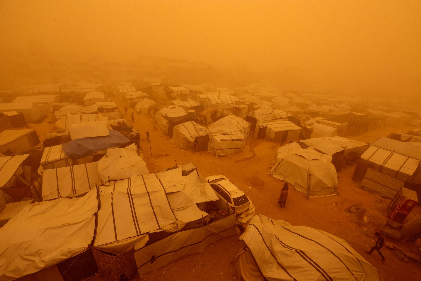 People walk amid a sandstorm in a tent camp, under an orange sky.