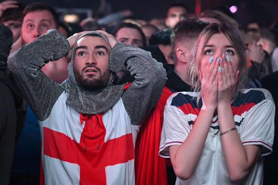 Two England fans look up at a screen and react sadly.