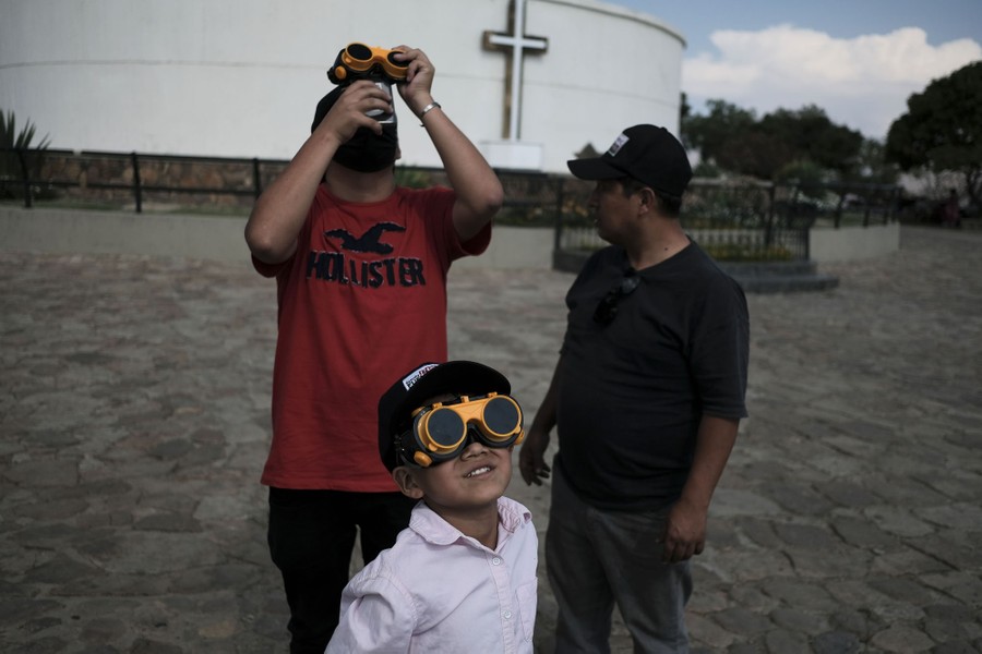 Three people stand together in a plaza for the eclipse. Two of them use welding goggles to look at the sky.