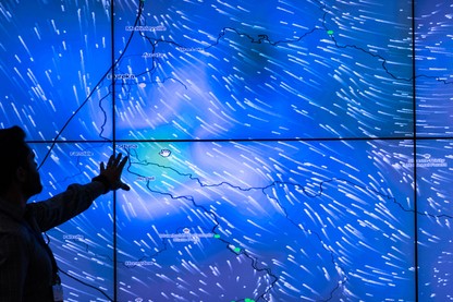 A meteorologist points to a big blue screen displaying wind data from hundreds of weather stations