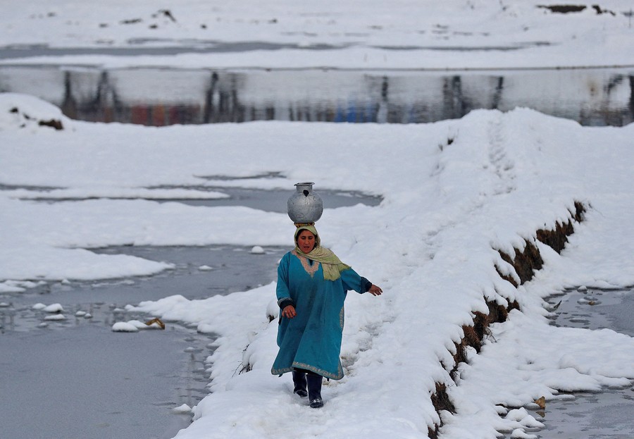 A woman carrying a water pitcher on her head walks through a snow-covered field.