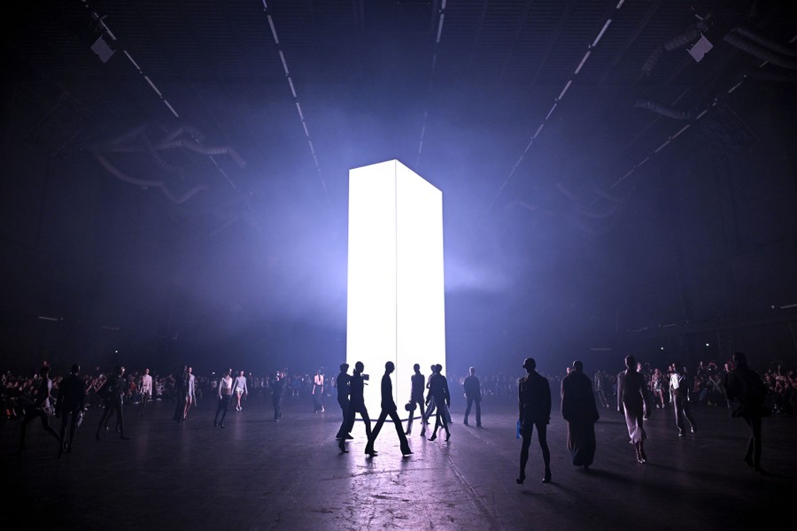 Models walk across an open space in a large dark room illuminated by a single tall light box in its center.