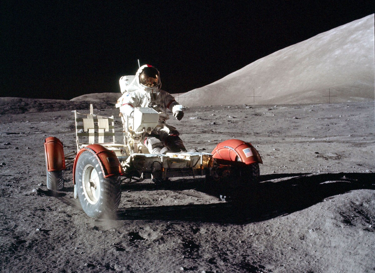 An astronaut drives a lunar rover across the surface of the moon toward the photographer.