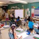 First-grade students are seated in a classroom listening to their teacher.