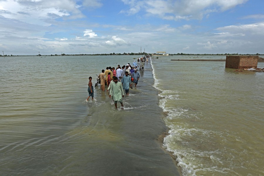A long line of people wade on a path through a flooded plain as water flows over a shallow ledge along the path.