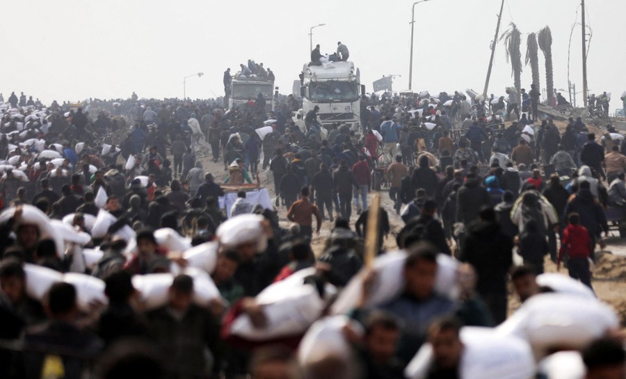 A large crowd gathers near two trucks, with many people carrying away bags of flour.