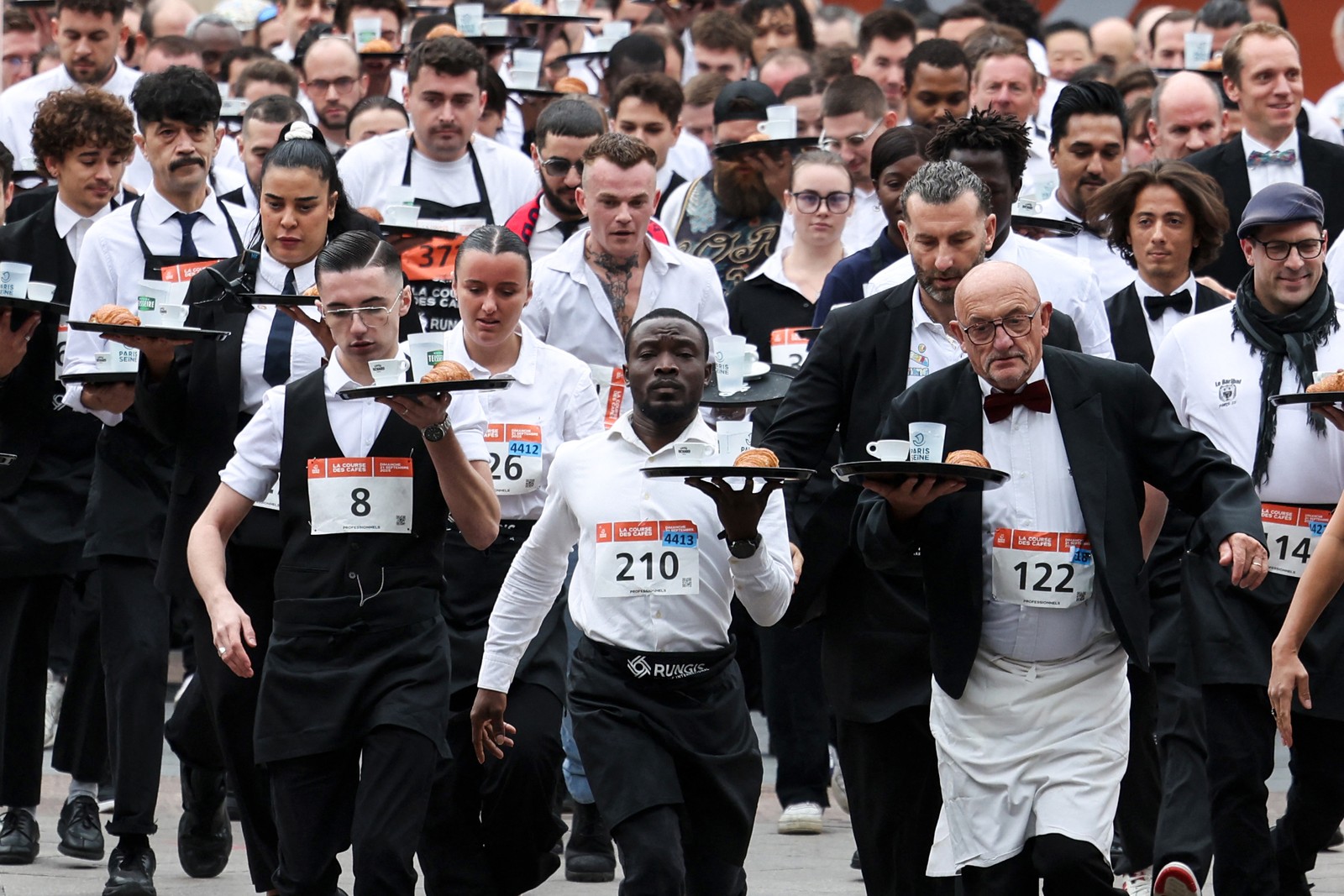 A group of waiters and waitresses, dressed in their work outfits, run a race while carrying a tray.